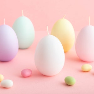 May include: Five pastel colored egg-shaped candles on a pink background with small pastel colored candy scattered around them.