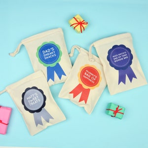 May include: Cream-colored drawstring bags with colorful ribbon designs and text. The bags read "Dad's Sneaky Snacks," "Mummy's Bag of Happiness," "Matt's Sweet Treats," and "Miss Smith's End of Term Goodie Bag." Small wrapped gifts are scattered around.