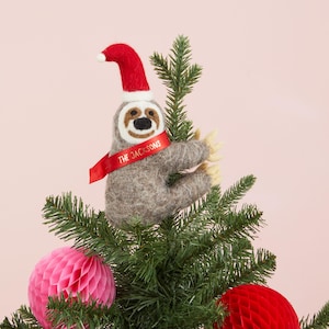 May include: A gray felt sloth Christmas tree topper wearing a red Santa hat and a red ribbon that says "THE JACKSONS". The sloth is hanging on a green Christmas tree with pink and red honeycomb ornaments.