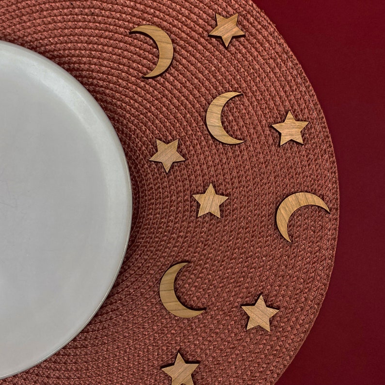 May include: A brown woven placemat with wooden crescent moons and stars scattered across the surface. A white ceramic plate is placed on the left side of the placemat.
