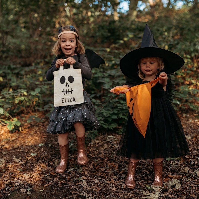 Pode incluir: Duas crian&ccedil;as vestidas com fantasias de Halloween. Uma crian&ccedil;a est&aacute; usando um traje de bruxa preto com um chap&eacute;u preto e segurando uma bolsa laranja em forma de ab&oacute;bora. A outra crian&ccedil;a est&aacute; usando um traje preto e cinza com uma capa de asa de morcego e segurando uma bolsa branca de doces ou travessuras com um design de caveira e o nome "Eliza" impresso nela.