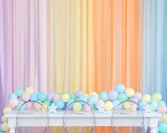 May include: A table setting with a pastel rainbow backdrop, featuring a white table with clear chairs, pastel balloons, and star-shaped decorations.