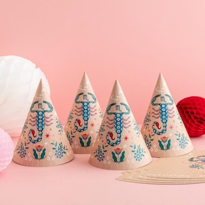 May include: Four beige party hats with a scorpion design in blue and red, surrounded by floral and star patterns. The hats are cone-shaped and decorated with the zodiac symbol. A few matching paper cutouts are visible.