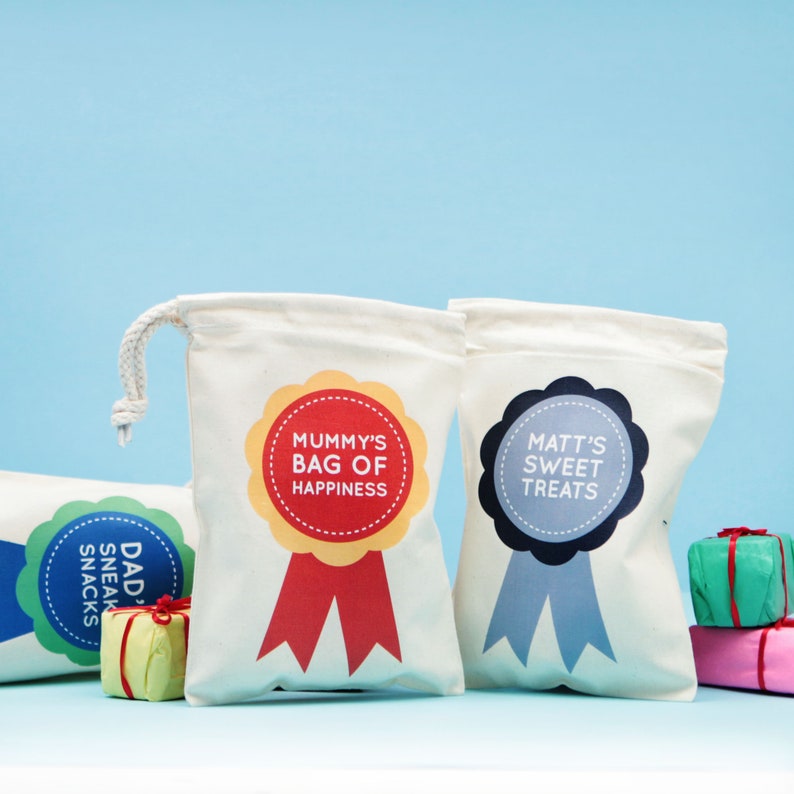 May include: Off-white canvas drawstring bags with colorful circular labels. One label reads "Mummy's Bag of Happiness" with red and yellow accents. Another says "Matt's Sweet Treats" with blue accents. A third says "Dad's Sneak Snacks".