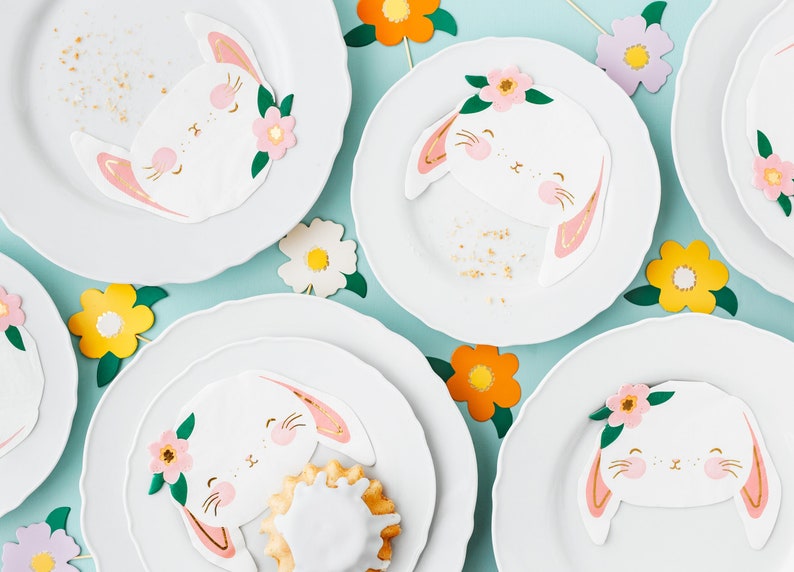 May include: White plates with scalloped edges are decorated with bunny-shaped napkins, featuring pink and gold accents, and floral embellishments. Colorful paper flowers in yellow, orange, and white add to the festive Easter theme.
