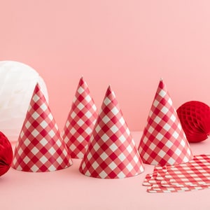 May include: Four red and white checkered party hats are arranged on a pink surface. The hats are cone-shaped and have a classic gingham pattern. Decorative red honeycomb balls and matching checkered paper are also present, suggesting a festive theme.