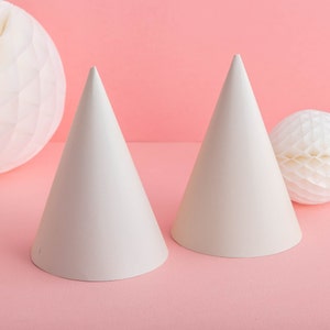 May include: Two white paper party hats on a pink background. The hats are pointed and have a smooth surface.