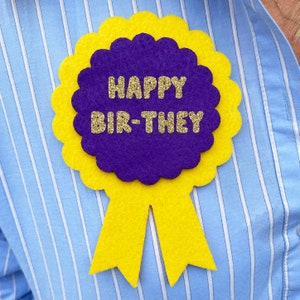 May include: A yellow and purple felt birthday badge with the text "Happy Bir-They".