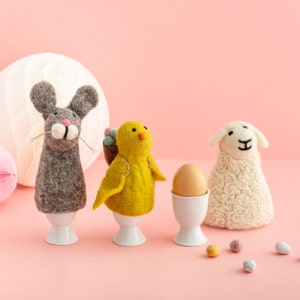 May include: Easter egg cups made of felt, including a grey bunny, a yellow chick with a basket, and a white sheep. A brown egg is in a white egg cup. Pastel speckled candy eggs are scattered on a pink background.