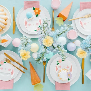 May include: An overhead shot of an Easter table setting with white plates, gold cutlery, and bunny-themed decorations. The table features cakes, pastel-colored eggs, and floral arrangements. Carrot-shaped and flower-shaped decorations add to the festive theme.