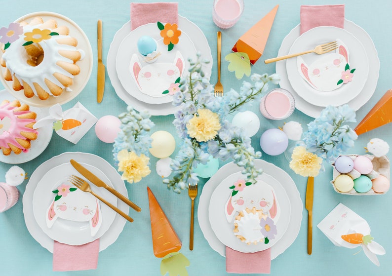 May include: An overhead shot of an Easter table setting with white plates, gold cutlery, and bunny-themed decorations. The table features cakes, pastel-colored eggs, and floral arrangements. Carrot-shaped and flower-shaped decorations add to the festive theme.