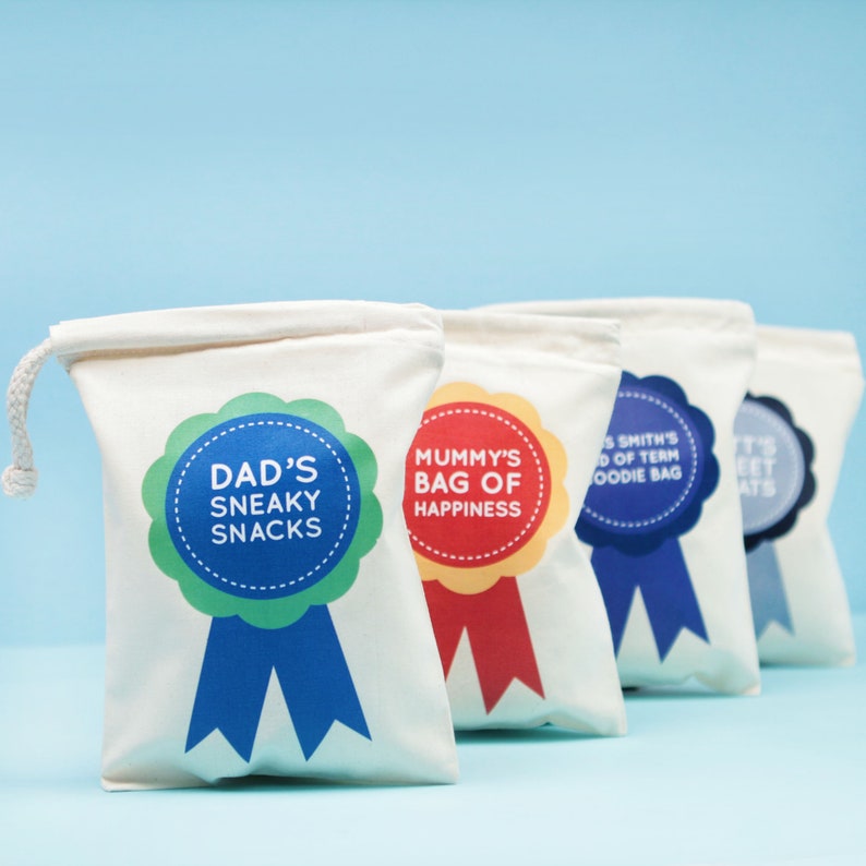 May include: Four white canvas drawstring bags with colorful ribbon-shaped labels. The labels read "Dad's Sneaky Snacks," "Mummy's Bag of Happiness," "Smith's End of Term Foodie Bag," and "Sweet Treats."