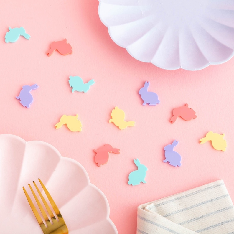 Pode incluir: Um fundo rosa com um prato rosa e um prato branco. H&aacute; formas de coelhos pastel espalhadas ao redor dos pratos. Um garfo dourado est&aacute; no prato rosa.