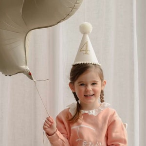 May include: A young child wearing a white party hat with a gold glitter number "4" on it. The child is smiling and holding a large, light brown balloon.