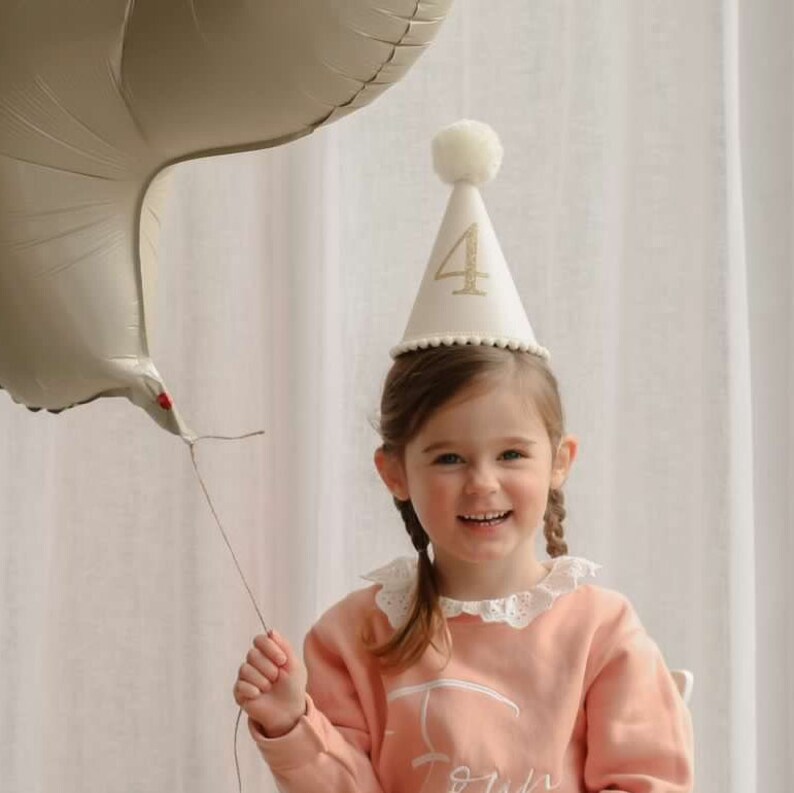 May include: A young child wearing a white party hat with a gold glitter number "4" on it. The child is smiling and holding a large, light brown balloon.