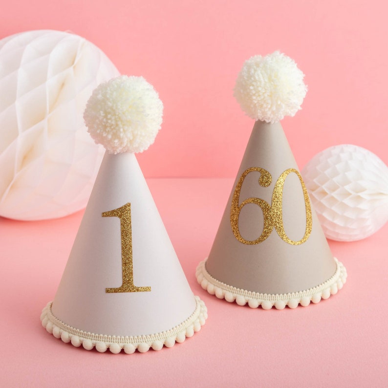 May include: Two party hats with white pom poms on top. One hat is white with a gold glitter number "1" and the other is beige with a gold glitter number "60".
