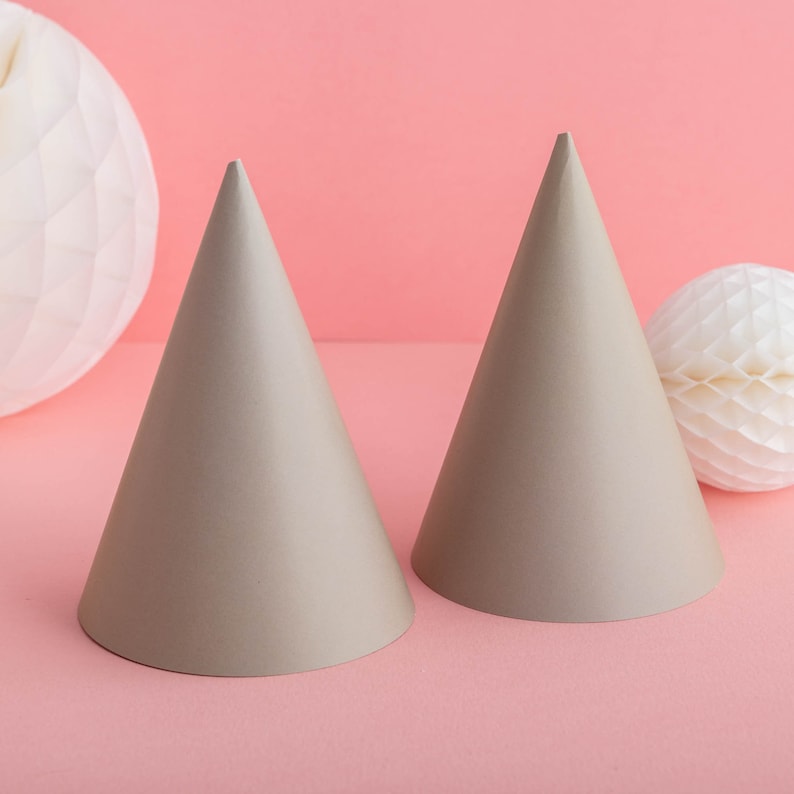 May include: Two light brown party hats on a pink background. The hats are made of paper and have a pointed tip.