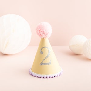 May include: A yellow party hat with a pink pom-pom on top and a silver glitter number "2" on the front. The hat has a purple and white trim.