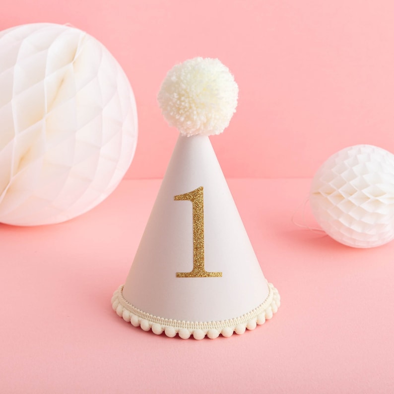 May include: A white party hat with a white pom-pom on top and a gold glitter number "1" on the front. The hat is decorated with a white trim around the bottom.