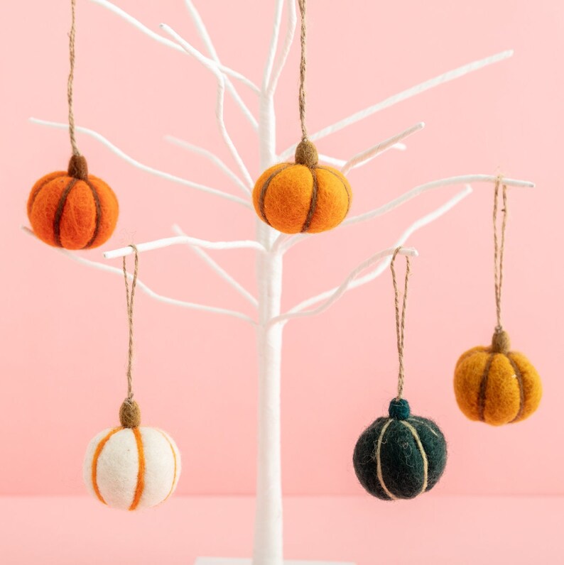 May include: Five felt pumpkin ornaments hanging from a white branch tree. The pumpkins are in various colors, including orange, white, and green.