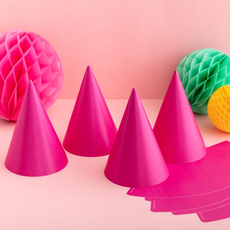 May include: Four pink party hats arranged on a pink background with two honeycomb balls, one green and one yellow.