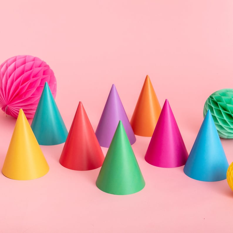May include: A set of nine colorful party hats in various shades of red, orange, yellow, green, blue, purple, and pink. The hats are arranged in a row on a pink background.