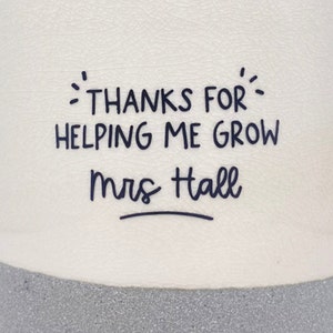 May include: White ceramic planter with a grey base. The planter has black text that reads "Thanks for helping me grow Mrs. Hall" with a line under the name.