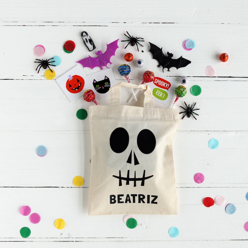 Pode incluir: Uma sacola de lona branca com um rosto de caveira preto e o nome "BEATRIZ" impresso na frente. A sacola est&aacute; cheia de guloseimas com tema de Halloween, incluindo doces, adesivos e brinquedos.