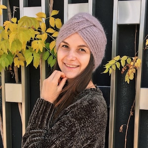 May include: A woman wearing a purple knitted turban hat with a knot on top. She is wearing a brown sweater and is smiling at the camera.