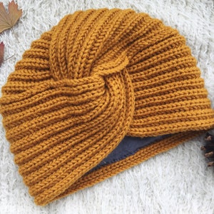 Könnte beinhalten: Ein turbanförmiger Strickhut in Senfgelb mit einem geknoteten Design. Der Hut ist aus weichem, strukturiertem Garn gefertigt.