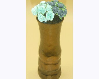 Puede incluir: Un jarrón de madera con un acabado marrón claro y una abertura ligeramente acampanada. El jarrón está lleno de un ramo de flores de papel, incluyendo rosas azul claro y gris oscuro.