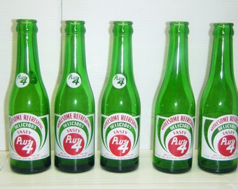 May include: Five green glass bottles with white labels and red lettering. The labels read "Wholesome Refreshing Delicious Tasty Plus 4".