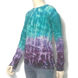 May include: A long-sleeved tie-dye shirt in shades of teal, purple, and black. The shirt has a crew neck and a gradient effect, transitioning from teal at the top to purple and black at the bottom. The sleeves are long and fitted.