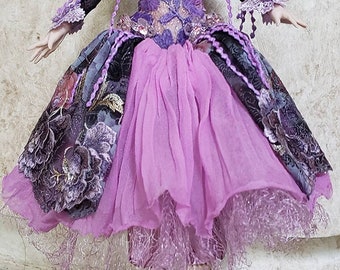 May include: A porcelain doll with brown hair and blue eyes wearing a purple and black dress with lace details. The doll is sitting on a glass pedestal.