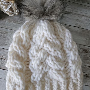 May include: White knitted beanie with a large, fluffy black pom pom. The beanie has a cable knit pattern.