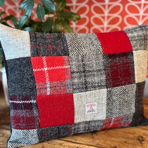 Peut inclure: Un coussin rectangulaire composé d'un assemblage de tissus en laine dans des tons de rouge, gris anthracite, gris clair et noir. Les motifs incluent des unis, des carreaux et des chevrons. Une petite étiquette Harris Tweed est cousue dessus.