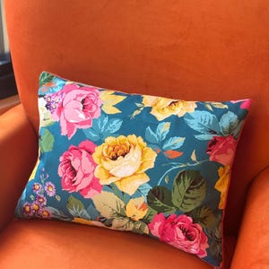 Puede incluir: Un cojín rectangular con fondo verde azulado y un vibrante estampado floral de grandes rosas rosas y amarillas, hojas verdes y pequeñas flores moradas. El cojín presenta una cremallera visible a lo largo del borde lateral y un borde naranja.