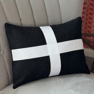 May include: Black and white throw pillow with a white cross design. The pillow is on a gray couch.