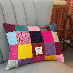 May include: A rectangular patchwork pillow with a variety of colorful squares in shades of pink, blue, yellow, and purple. The pillow is resting on a gray upholstered chair. A small label is visible on the pillow.