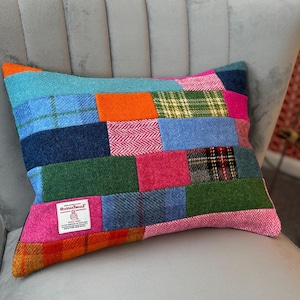 May include: Rectangular patchwork pillow with a variety of colorful tweed fabric squares. The pillow features a mix of blues, greens, pinks, and oranges. A white label with black text is visible on the front. The pillow is resting on a gray chair.