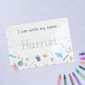 May include: White dry-erase board with the text "I can write my name" and the name "Harriet" written in dotted lines. The board is decorated with colourful school-themed illustrations.