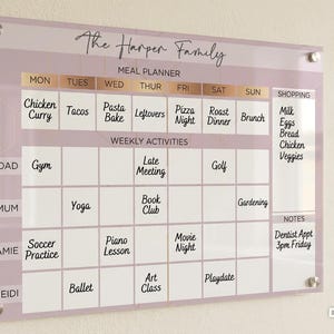 May include: A rectangular acrylic wall-mounted meal planner and activity calendar with a mauve and white geometric background. It features sections for The Harper Family, a weekly meal plan, daily activities for four people, a shopping list, and notes.