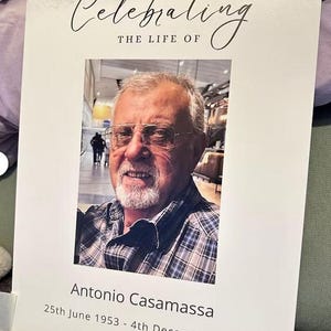 May include: A memorial card with the text "Celebrating The Life Of" above a portrait of a smiling older man wearing glasses and a plaid shirt. The name "Antonio Casamassa" and dates "25th June 1953 - 4th December" are printed below the photo.