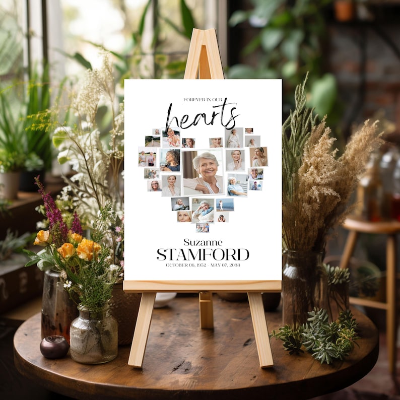 May include: A white memorial sign on a wooden easel, featuring a heart-shaped collage of photos. The sign reads "Forever in our hearts" and includes the name "Suzanne Stamford" and dates "October 06, 1952 - May 07, 2018".