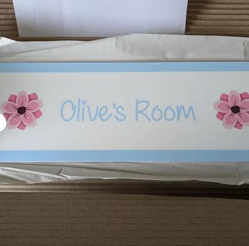 Puede incluir: Un letrero rectangular de madera con el texto "Olive's Room" en escritura azul claro. El letrero tiene un fondo blanco con un borde azul claro. Ilustraciones de flores rosas y rosa oscuro a cada lado del texto.