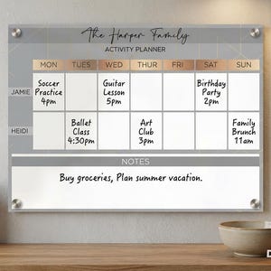 May include: A rectangular acrylic wall-mounted activity planner with silver hardware. The board features a gray and white grid labeled The Harper Family, with columns for days of the week and rows for names, plus a section for notes at the bottom.