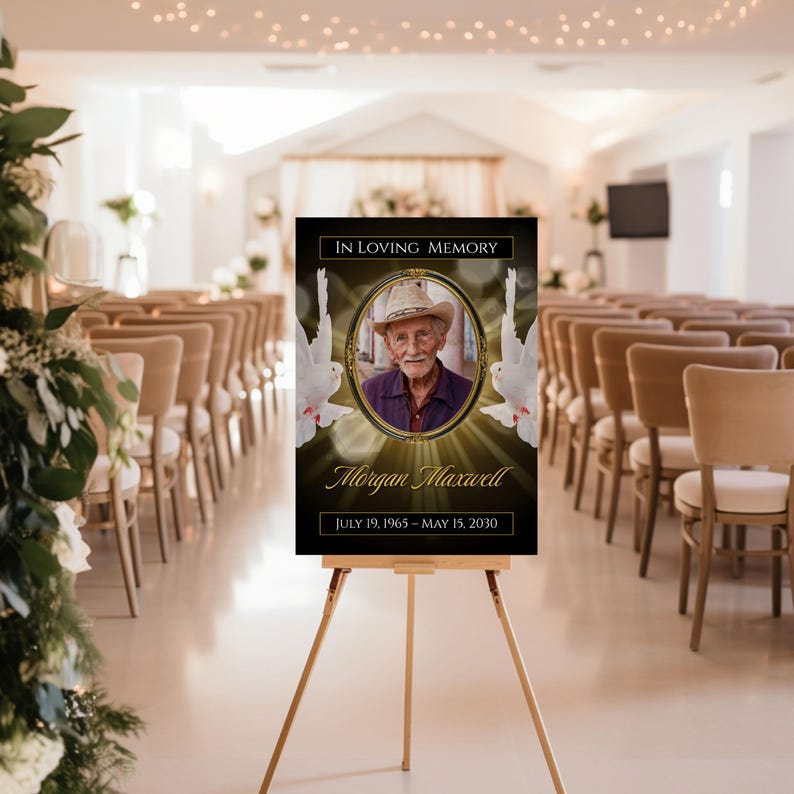 May include: A memorial sign on a wooden easel. The sign is black with gold accents and features a photo of a man in a hat, the text "In Loving Memory," and the dates July 19, 1965 - May 15, 2030. Two white doves flank the photo.