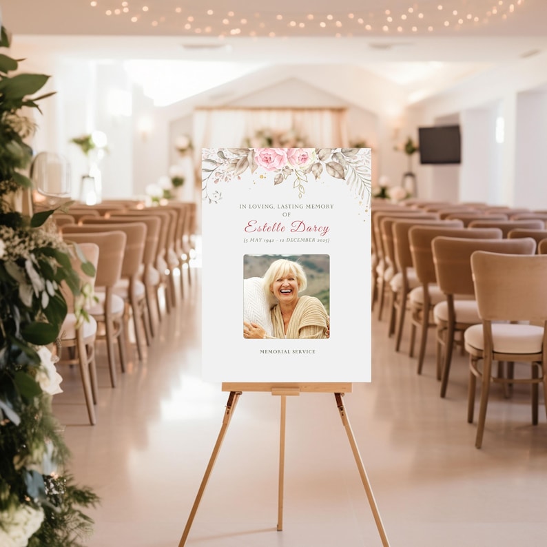 May include: A white memorial sign on a wooden easel, featuring a photo of a smiling woman and text. The sign is decorated with pink roses and foliage. The sign reads "In Loving, Lasting Memory of Estelle Darcy".