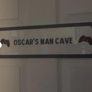 Puede incluir: Un letrero rectangular con el texto "OSCAR'S MAN CAVE" en letras negras. El letrero tiene un borde negro y presenta un gr&aacute;fico de mando de videojuegos a cada lado. El letrero est&aacute; montado en una puerta blanca.