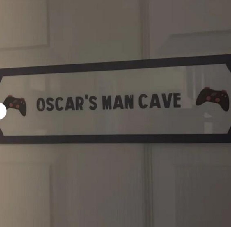 Puede incluir: Un letrero rectangular con el texto "OSCAR'S MAN CAVE" en letras negras. El letrero tiene un borde negro y presenta un gr&aacute;fico de mando de videojuegos a cada lado. El letrero est&aacute; montado en una puerta blanca.